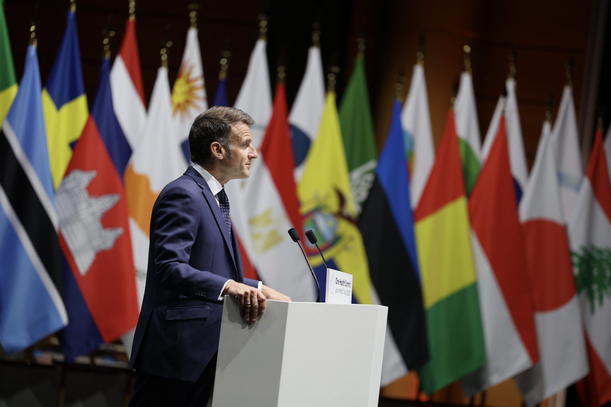 Photo of Presidenbt Macron in front of various country flag on the stage of the One Heanth Summit